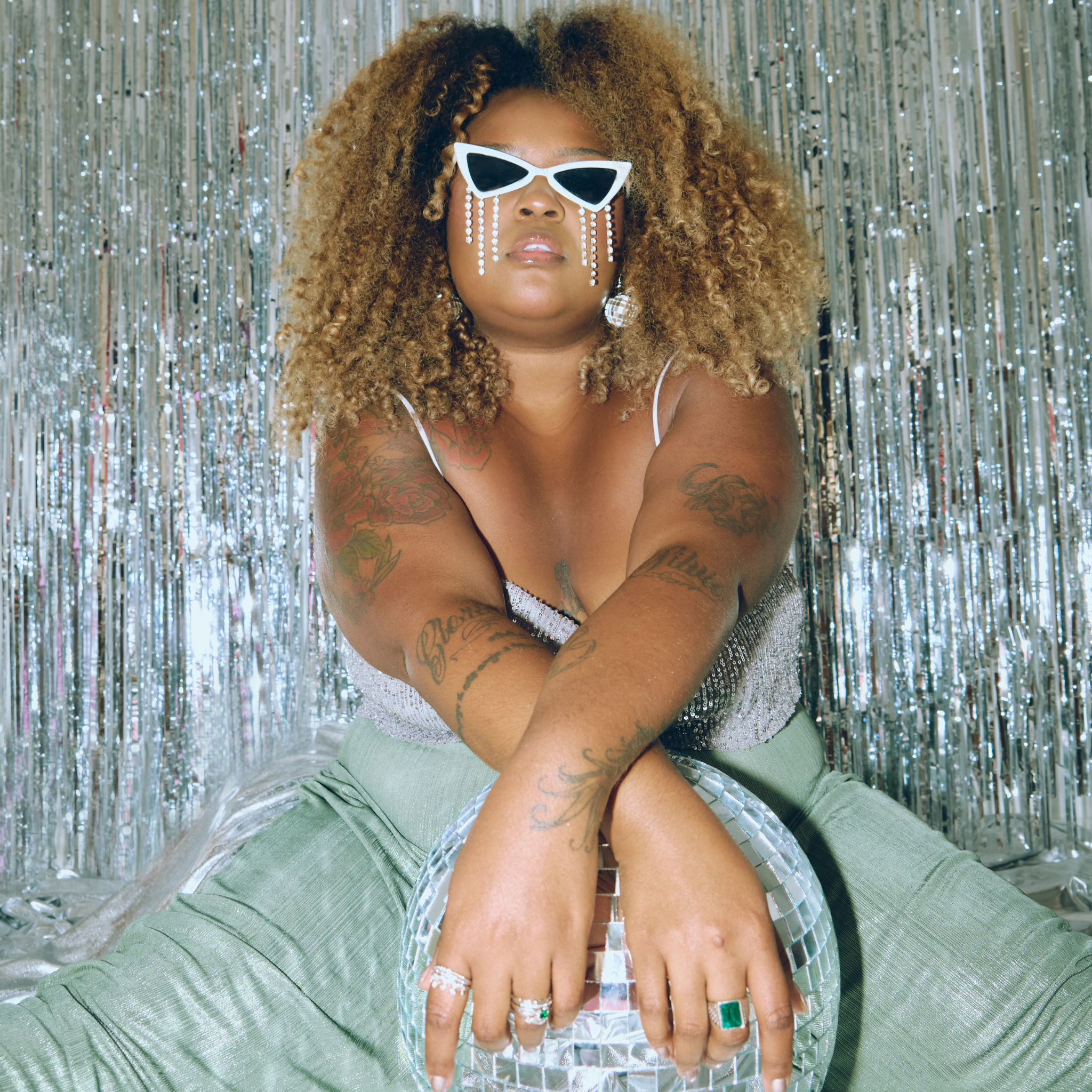 le bleu bodycare_Woman with tattoos wearing sunglasses and a green outfit against a silver tinsel background